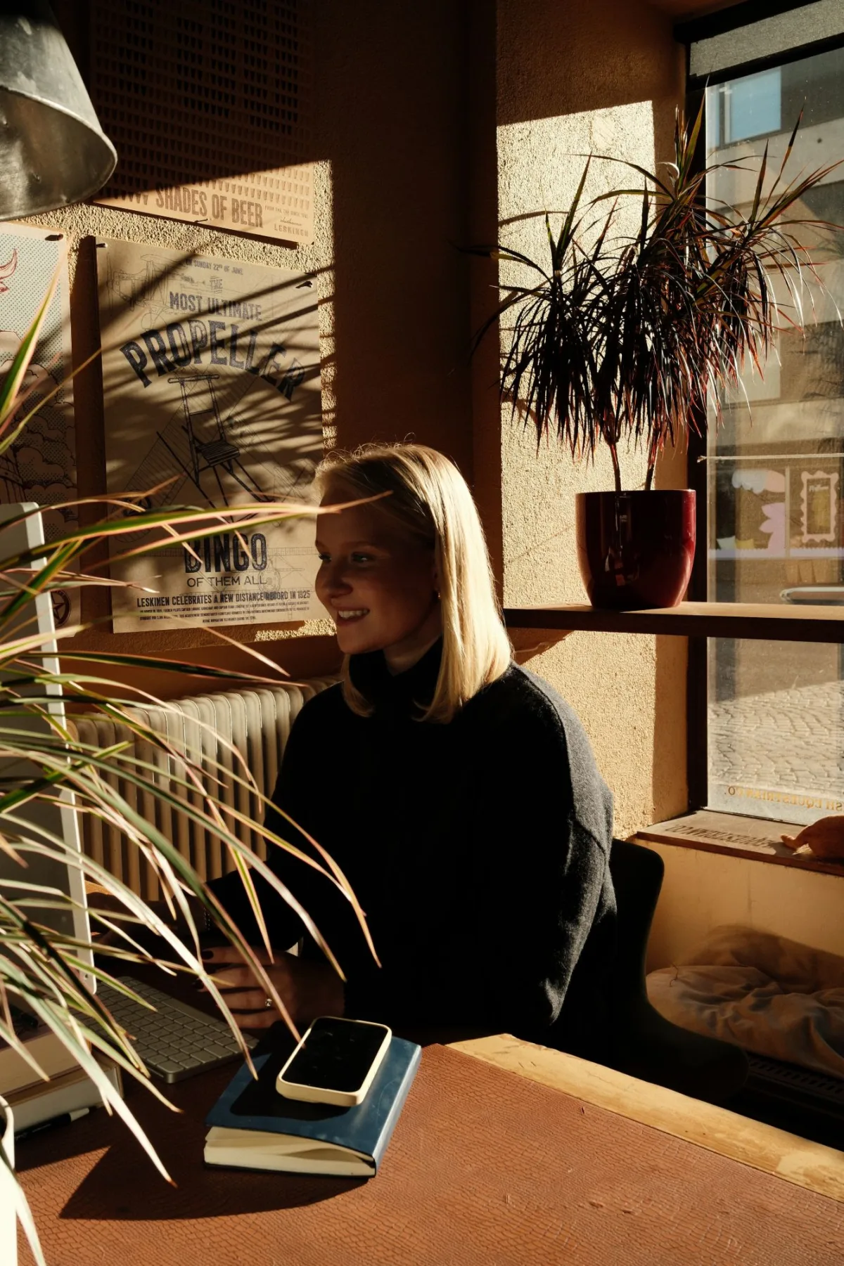 Iines Mujunen työpöydän ääressä hymyillen, ikkunasta lankeava iltapäivän valo huoneessa