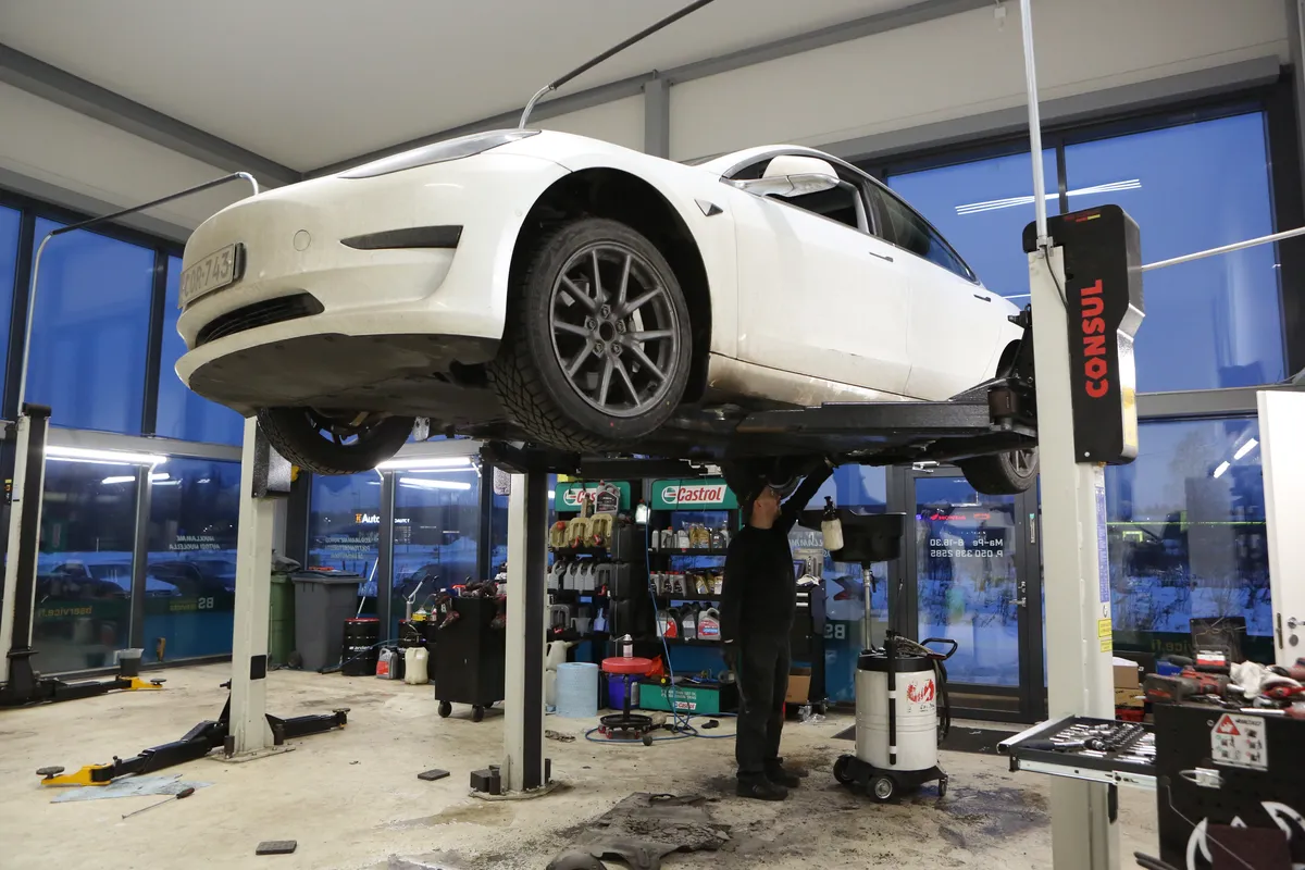 Auto nosturilla korjaamohallissa, asentaja työskentelee auton alla